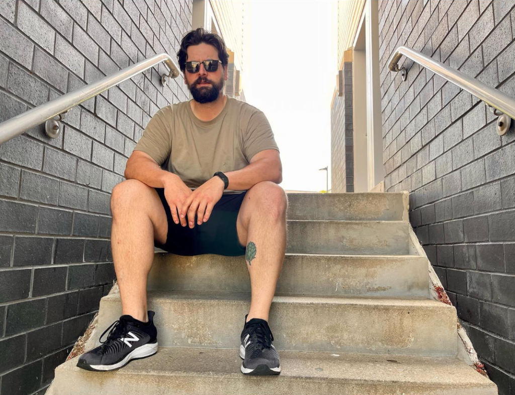 A man with dark hair and a dark beard and sunglasses sitting on steps outside with brick walls on either side of him.