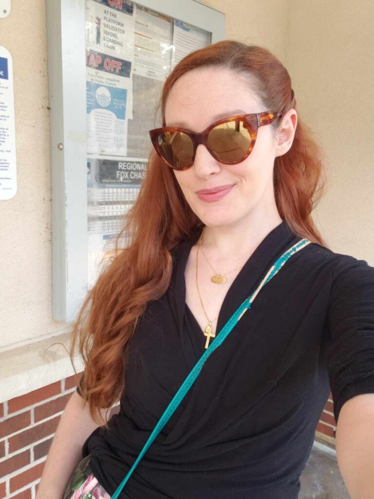A woman with long red hair and amber sunglasses. She is wearing a black shirt with a teal purse strap diagonally across it.