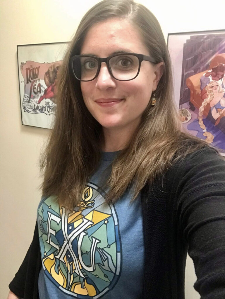 A woman with medium length brown hair and glasses. She is wearing a blue "Exandria Unlimited" T-shirt with a black cardigan.