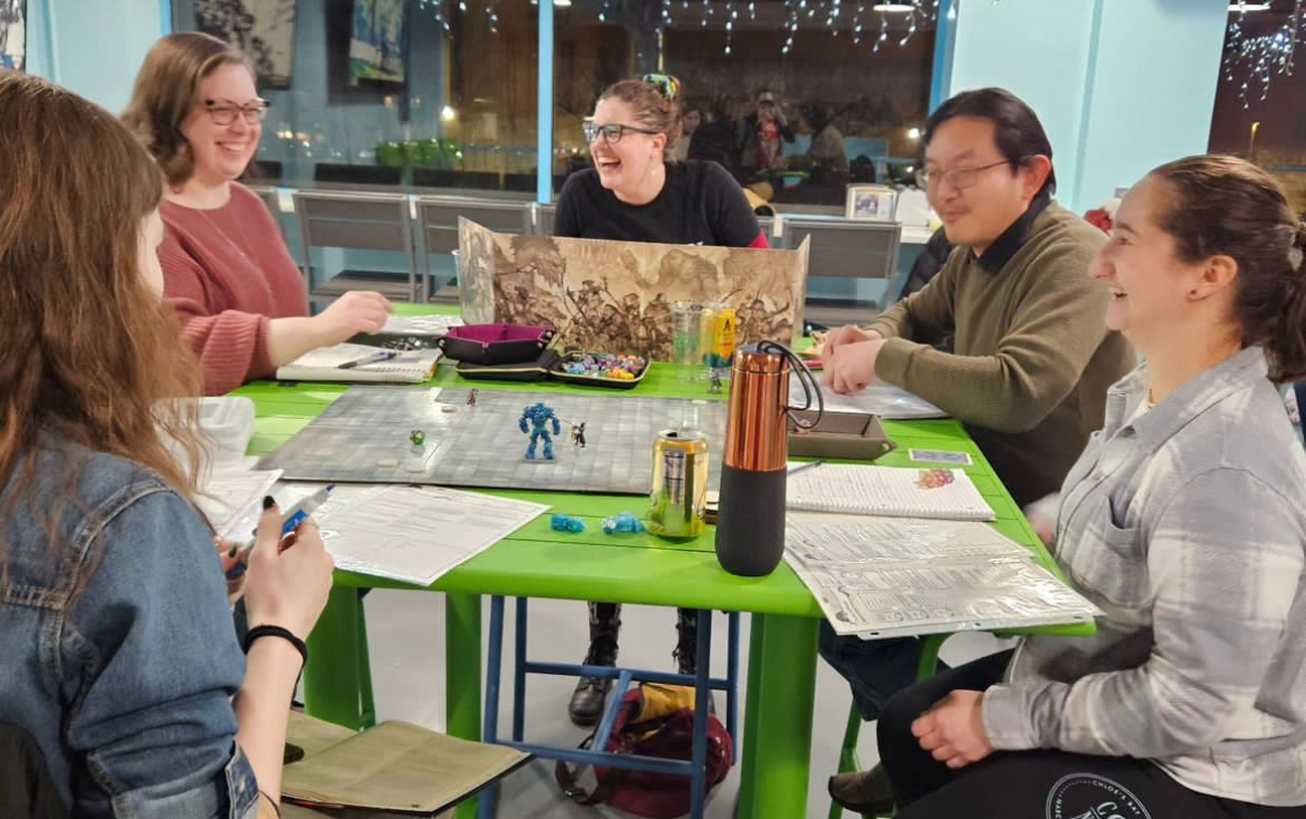 Female Dungeon Master sitting behind a DM screen. Other players surrounding her at the table.