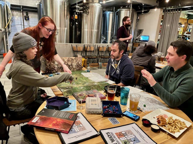 A photo from The Ways Restaurant & Brewery. It features a red haired woman acting as a dungeon master leading a party of adventurers at the table.