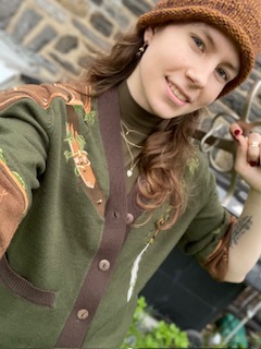 Cece - A female DM wearing a green sweater and brown cap, holding a sword over her shoulder.